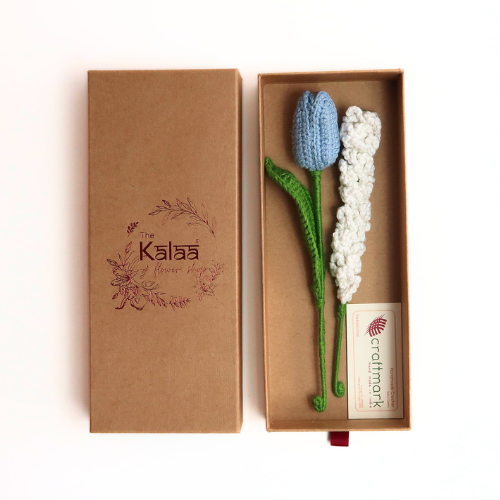 A gift box with a floral arrangement, primarily white and blue flowers, and a green knitted object resembling a stem, all presented on a brown cardboard backing with 'KALA'A' and 'craft-a-gift' logos.